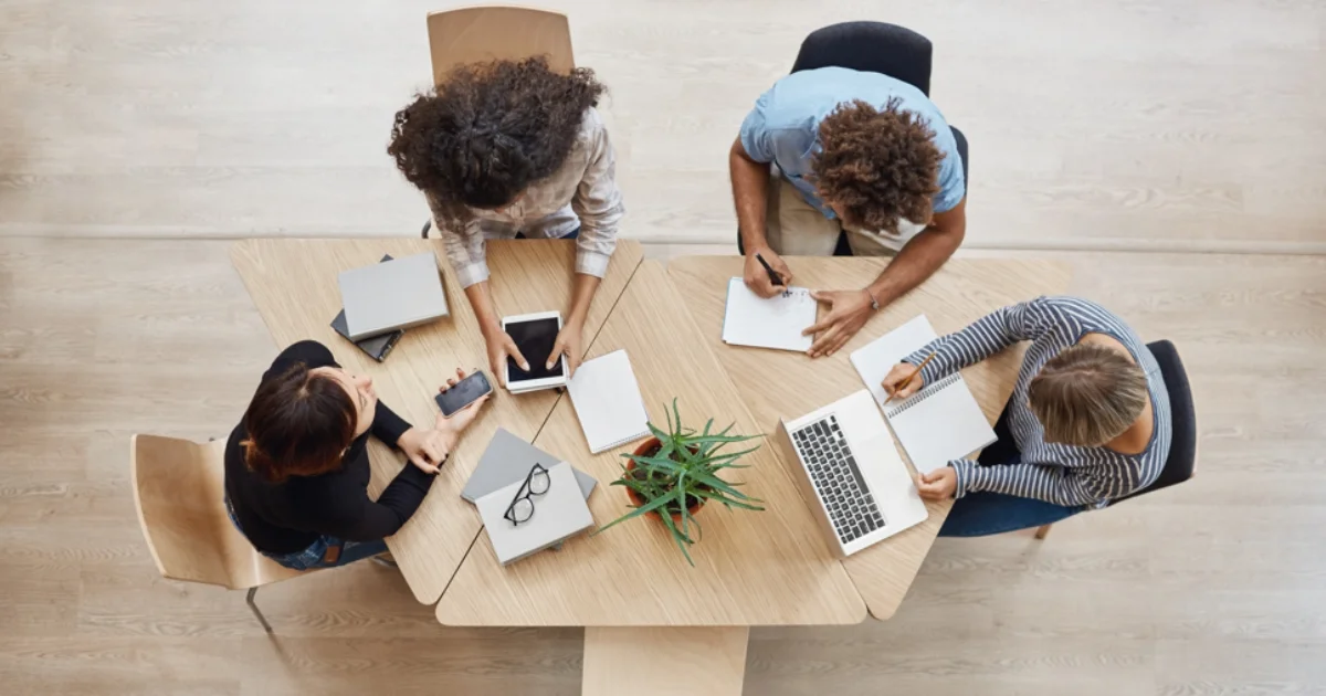 Overhead view of a diverse team working collaboratively at a modern wooden table with notebooks, laptops, and digital devices.